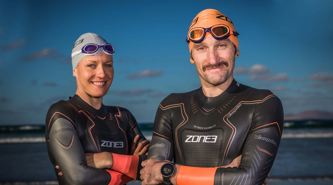 Two Zone3 athletes in wetsuits smile for a photo on the beach with the ocean and blue sky behind.