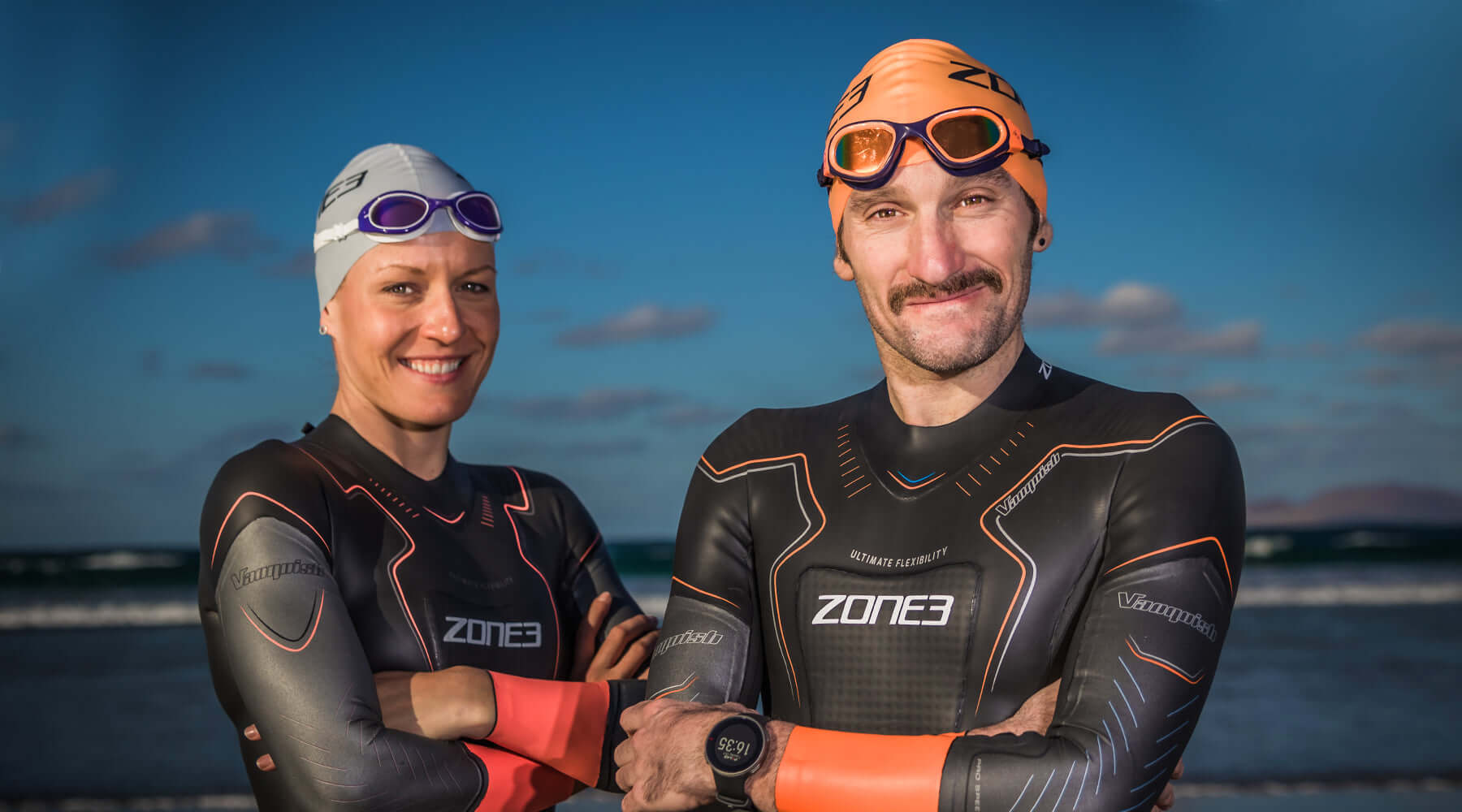 Two Zone3 athletes in wetsuits smile for a photo on the beach with the ocean and blue sky behind.