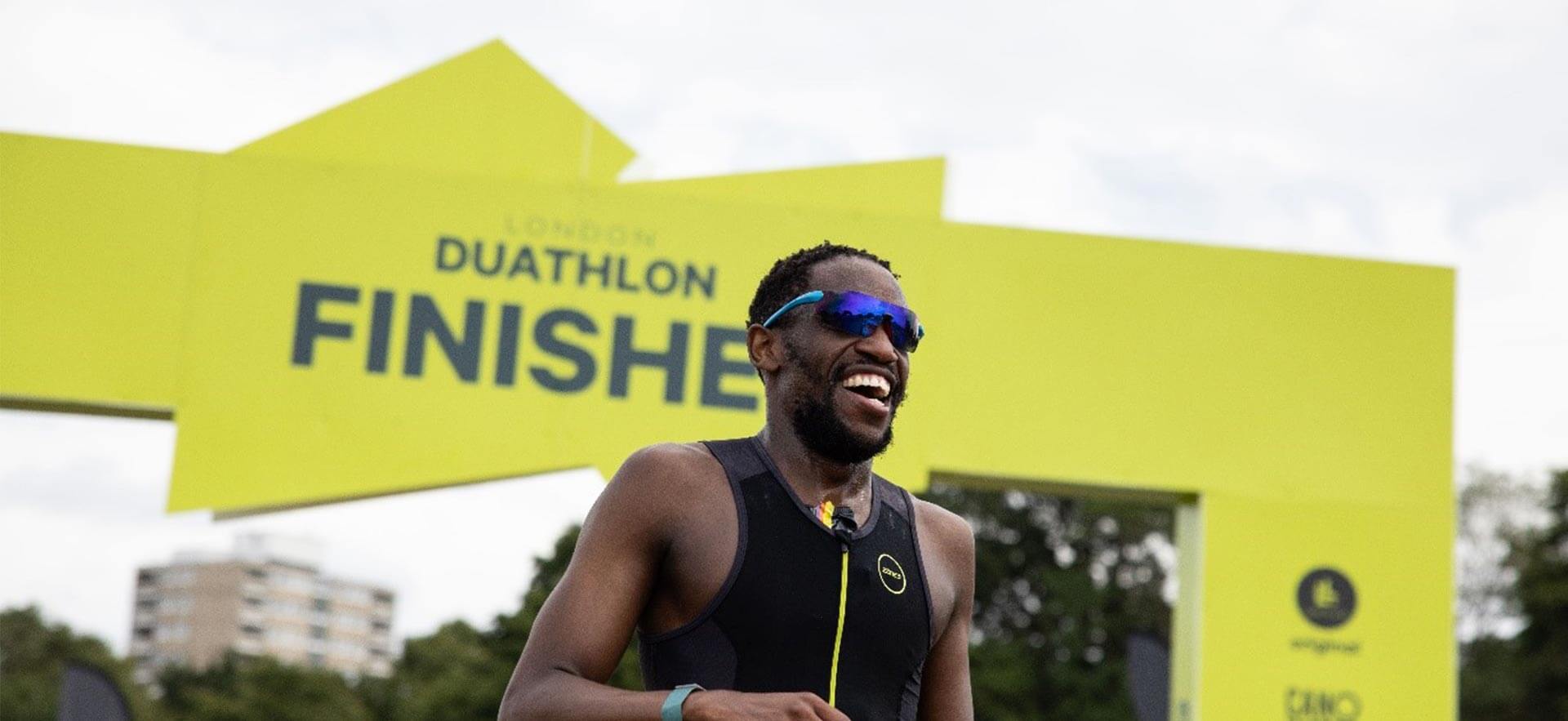 Smiling male athlete in a black sleeveless top and blue sunglasses crosses a bright yellow finish arch labeled DUATHLON FINISHES