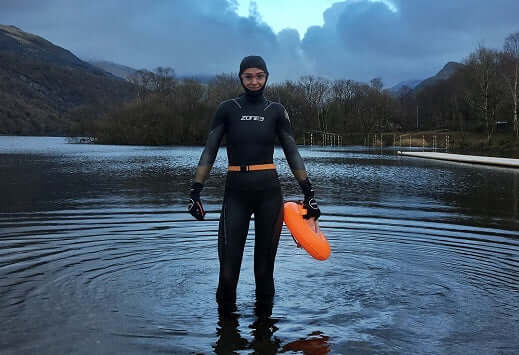 Swimmer in a black wetsuit stands in a tranquil lake holding an orange flotation buoy, mountains in the background.