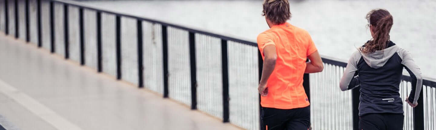 Two runners jog along a waterfront boardwalk with a metal railing, one in a bright orange shirt.