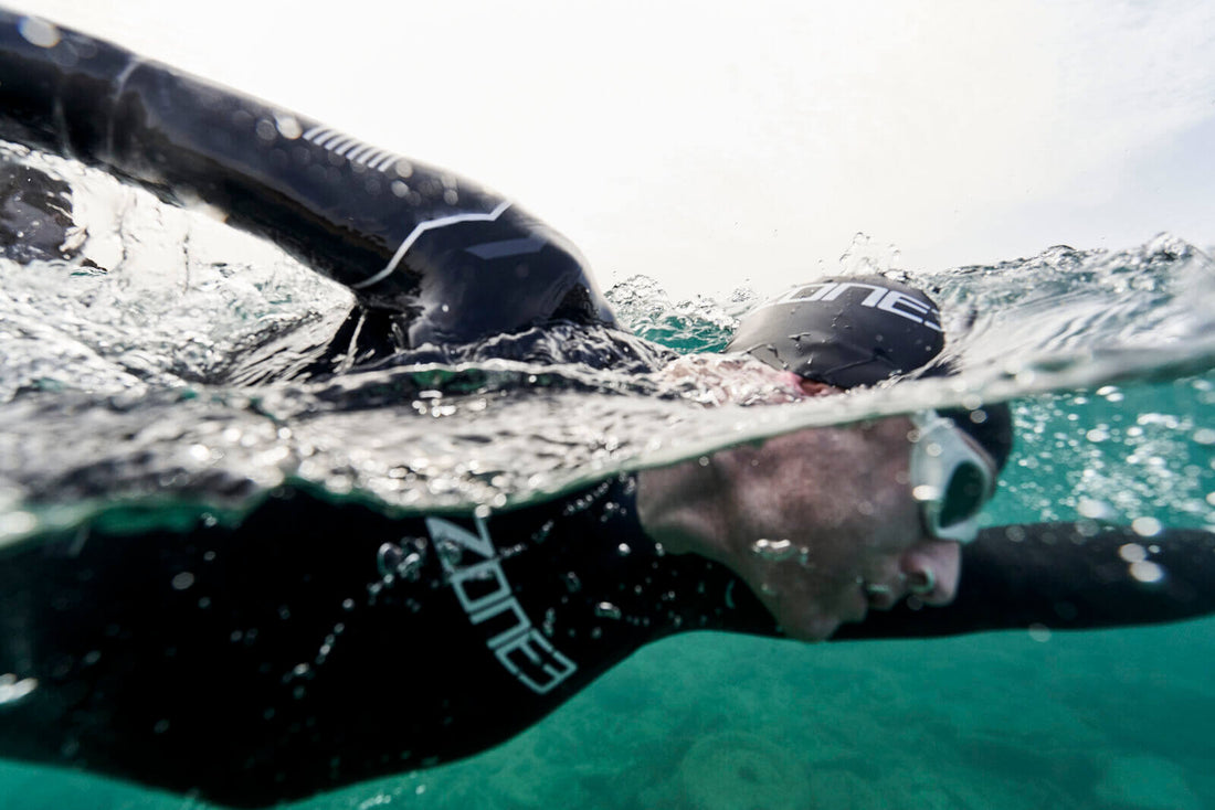Swimmer in a black Zone3 wetsuit with goggles glides through turquoise water near the surface