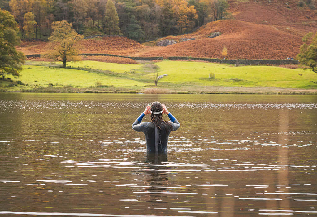 Swimmer in a wetsuit stands chest deep in a lake adjusting a swim cap as autumn hills rise in the background.