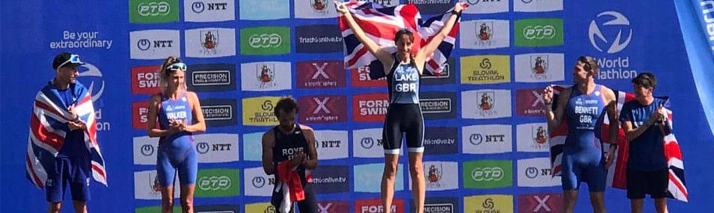 Athlete stands on a winners podium with a Union Jack behind, celebrating after a triathlon race.