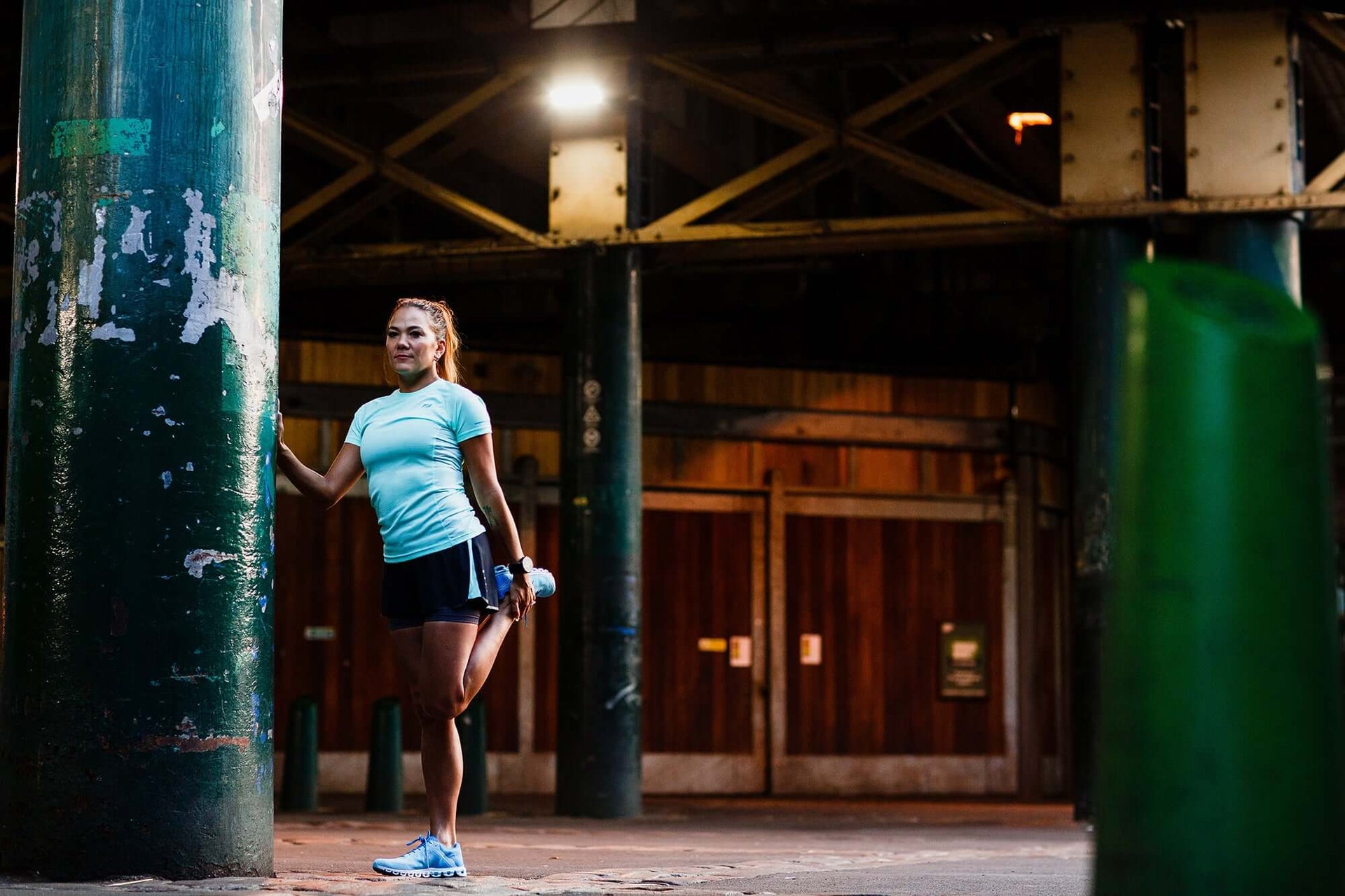 Female athlete in a light blue top and cap stands on concrete stairs inside an industrial gym.