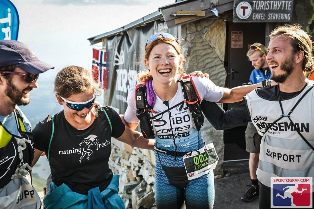 Smiling athletes celebrate at the Zone3 race finish area with a stone wall and banners in the background