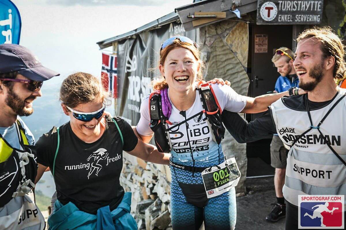 Smiling athletes celebrate at the Zone3 race finish area with a stone wall and banners in the background