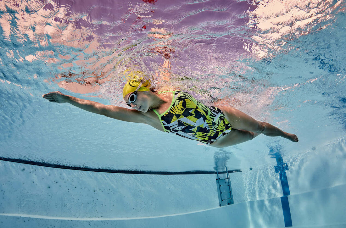 Swimmer in a geometric patterned swimsuit with a yellow cap and goggles gliding underwater.
