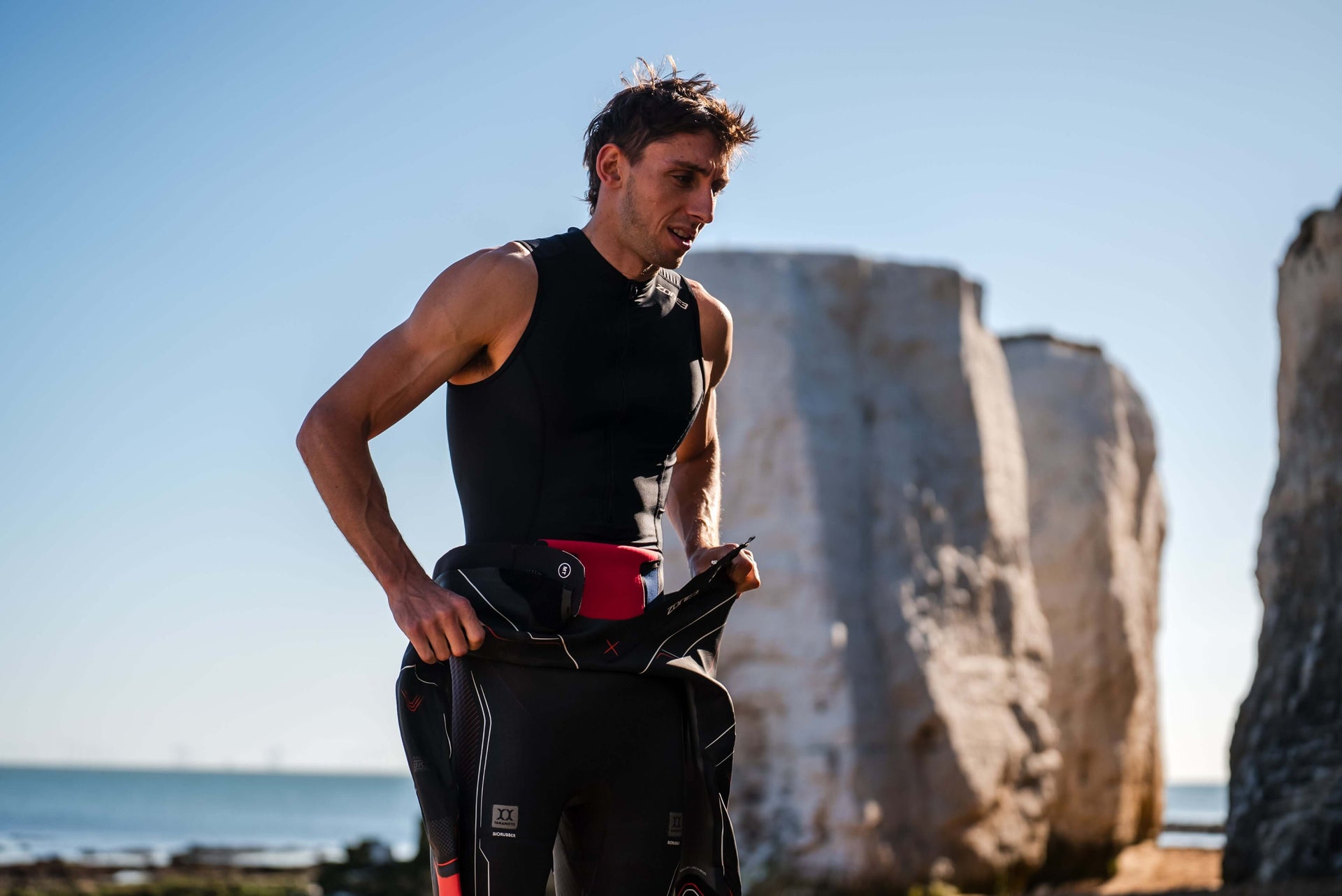 Athlete in a black wetsuit with a white cap stands by a rocky shoreline, wearing a red waterproof transition bag at the waist.