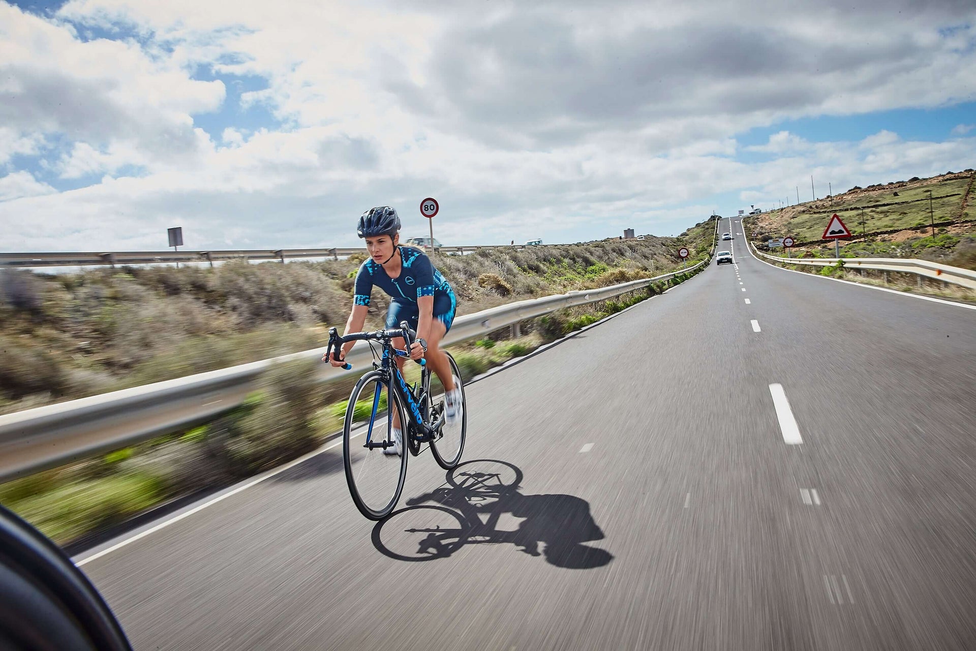 Cyclist in blue kit and helmet rides along a coastal road with an 80 sign in the distance