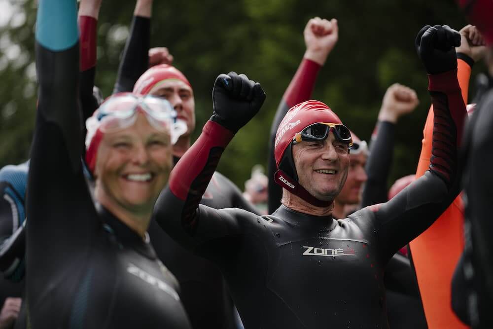 Swimmers in black wetsuits and red swim caps raise their arms in celebration at the Great North Swim event.