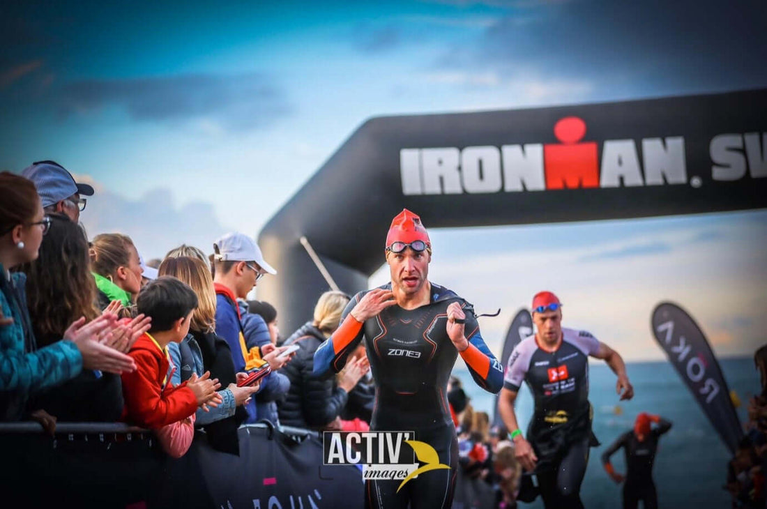 Triathlete in a red swim cap and wetsuit nears the finish under an Ironman banner as cheering spectators line the course.
