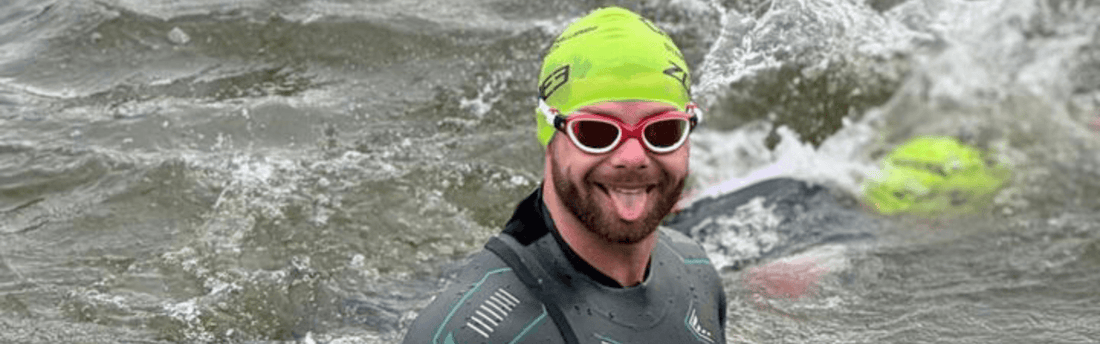 Triathlete wearing a neon green swim cap and red goggles smiles while swimming in choppy water.
