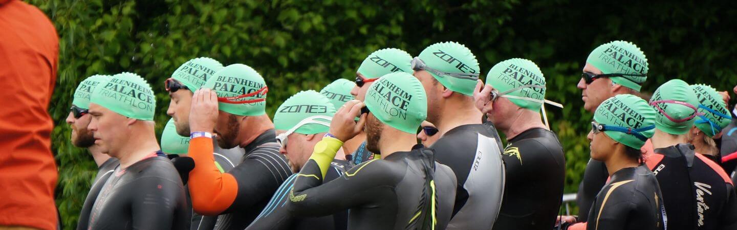 a group of people are at the swim start facing the water all with green swim caps on. 