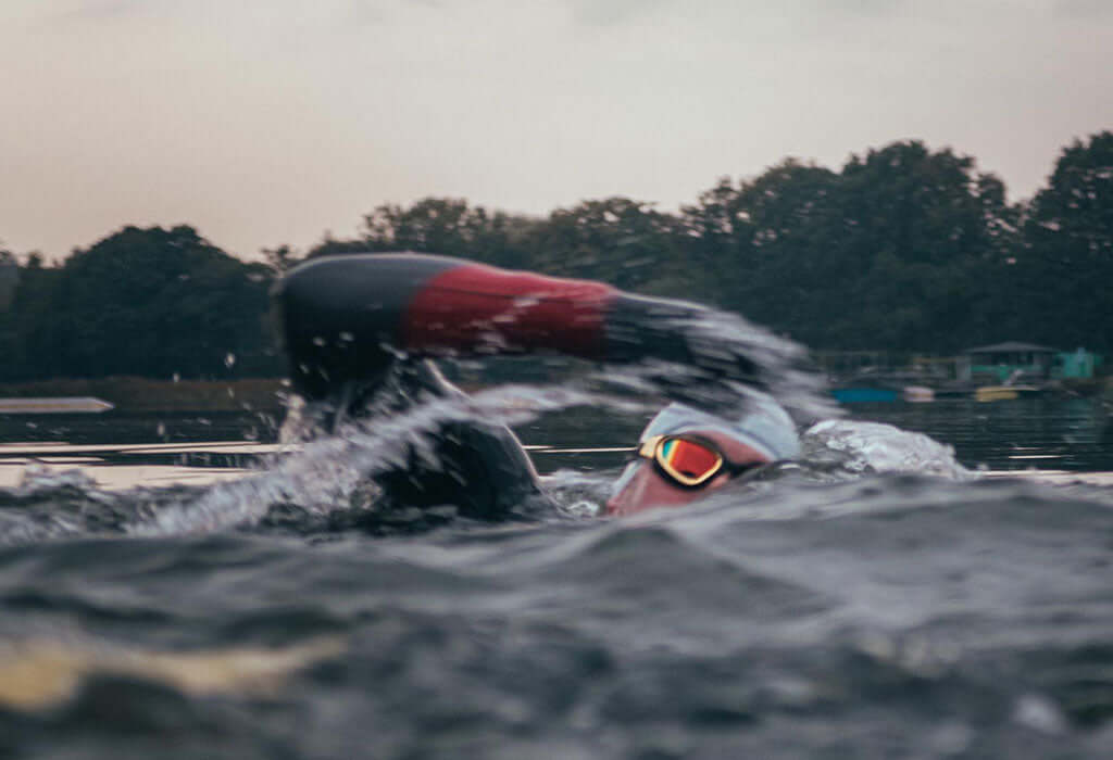 Open water swimmer wearing a cap and orange goggles glides through rippled lake water with trees along the shore.