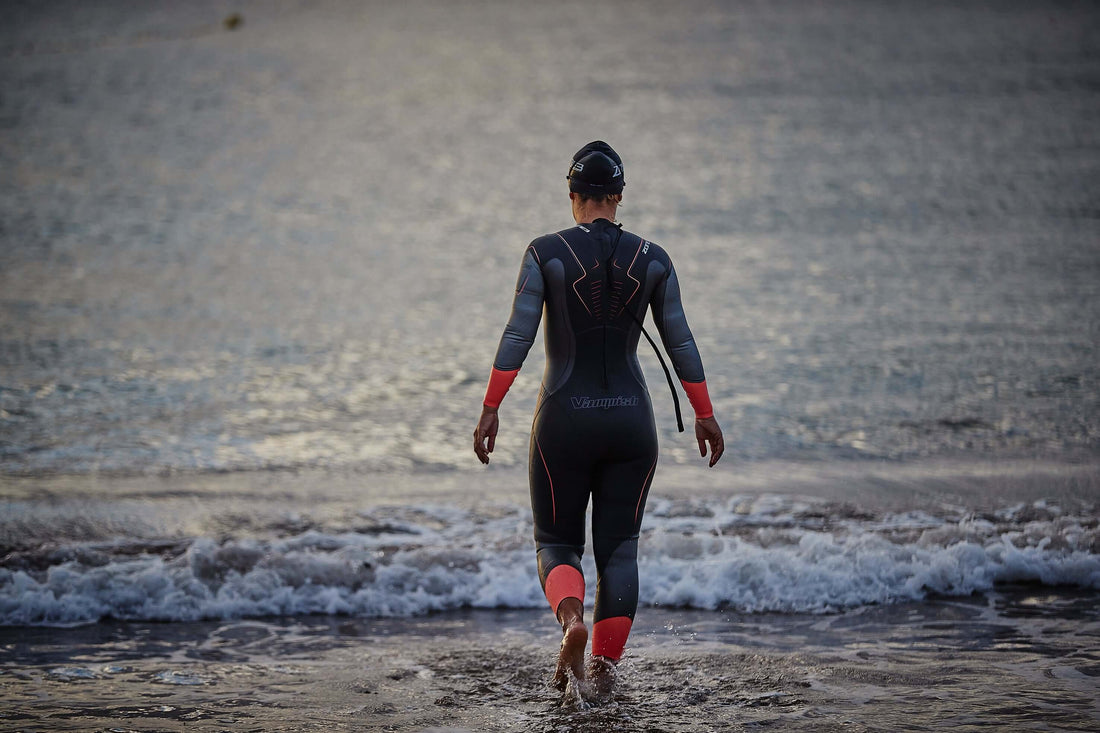 Triathlete in a black and red wetsuit walking toward the sea from a rocky shore, seen from behind