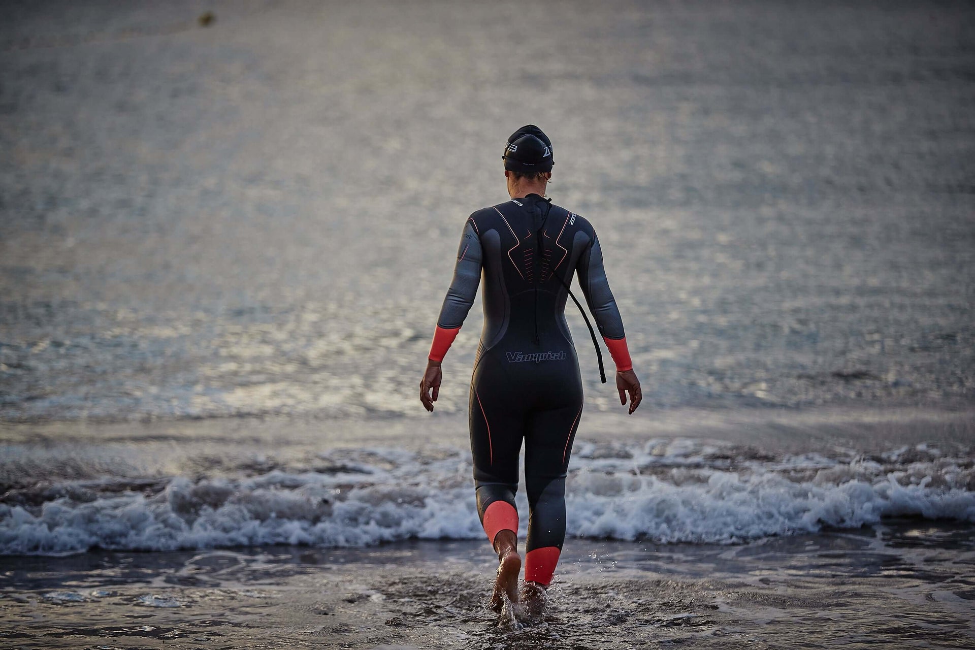 Triathlete in a black and red wetsuit walking toward the sea from a rocky shore, seen from behind
