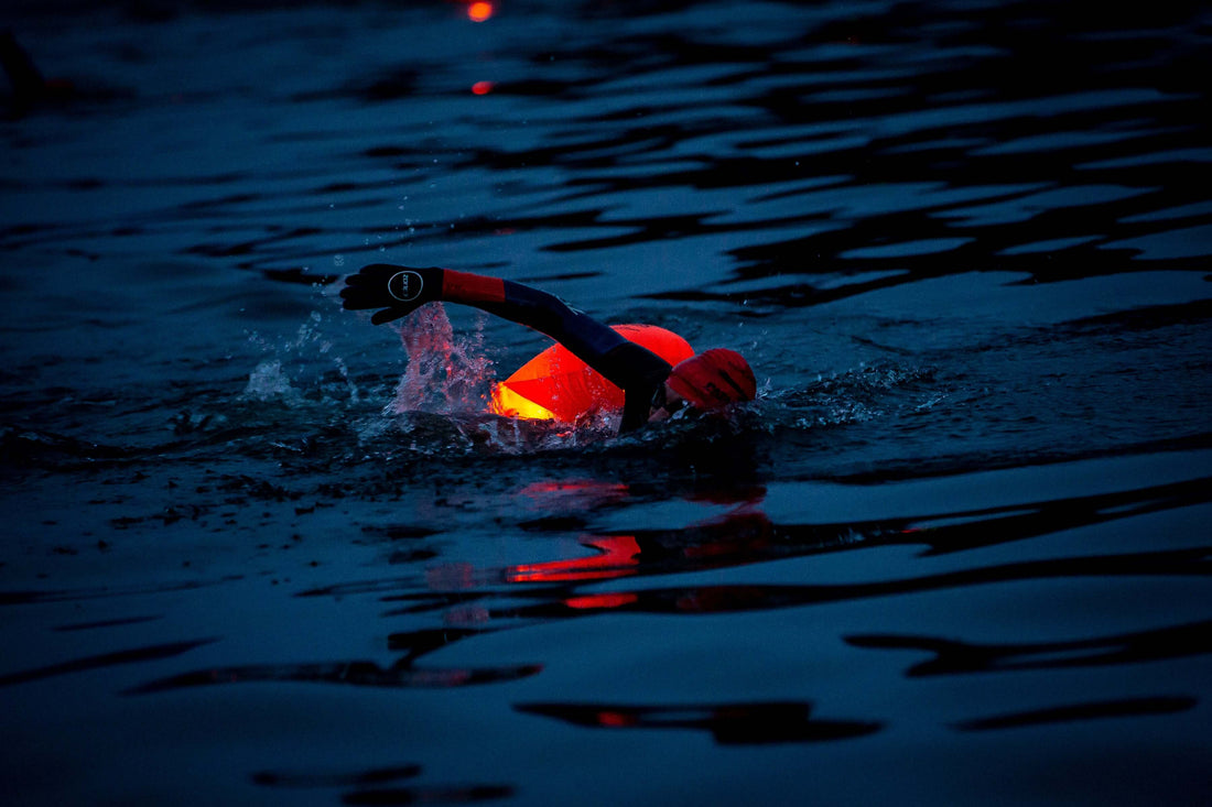 Swimmer wearing a red cap and gloves paddles through dark blue open water at dusk with a bright buoy nearby.