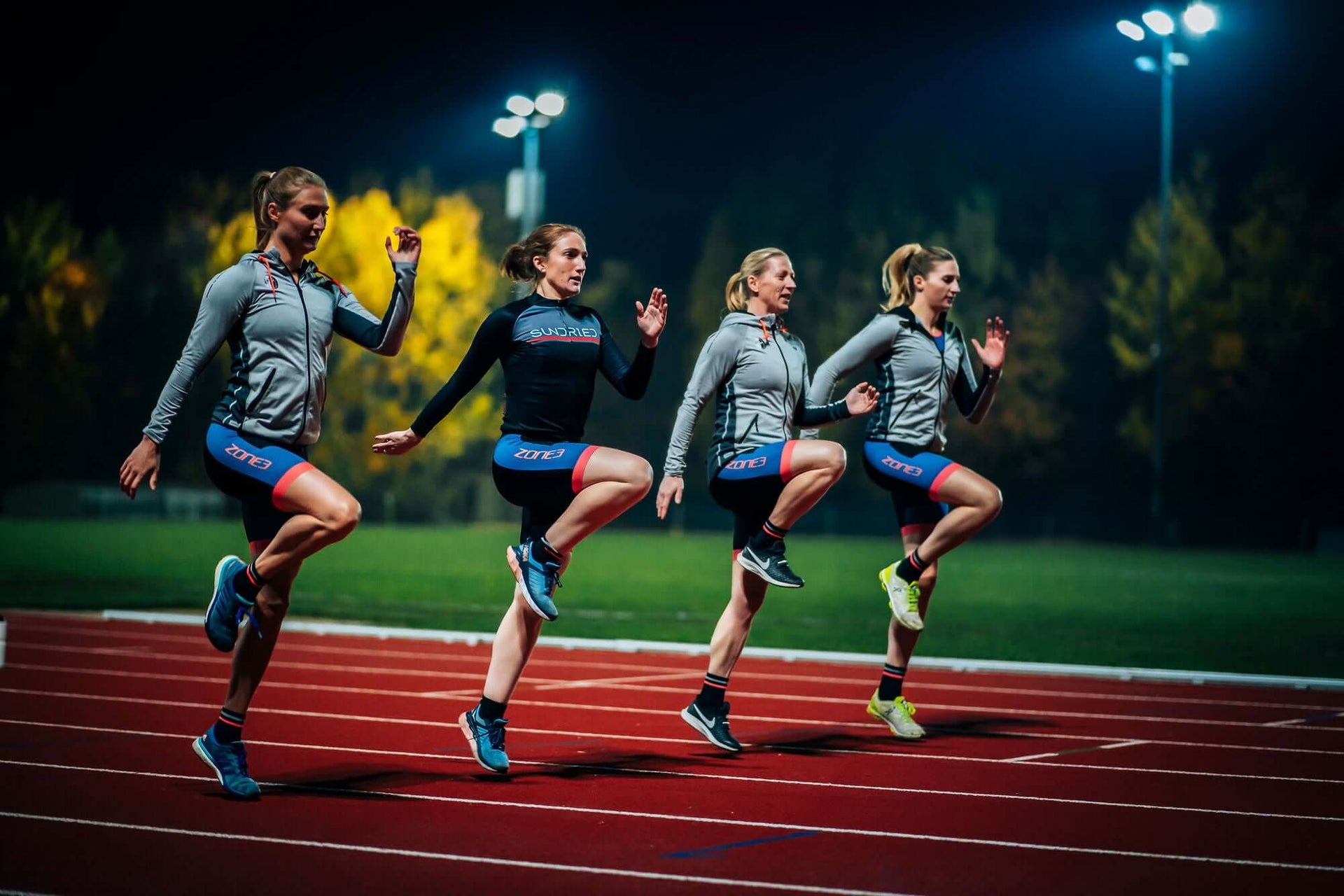 Four women runners sprint on a track at night under bright stadium lights in matching Zone3 gear