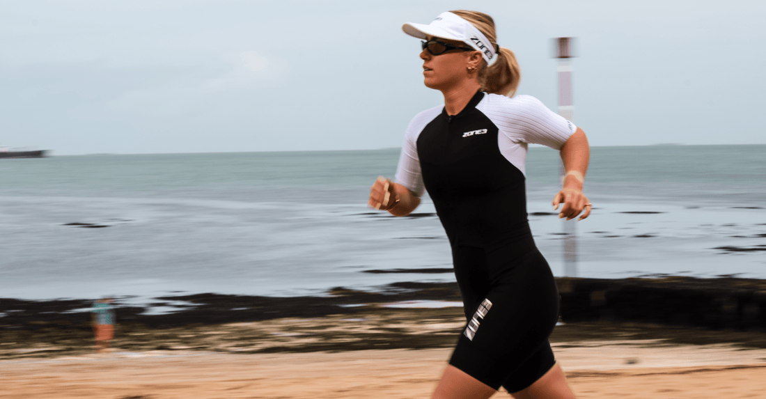 Female athlete in a black and white triathlon suit runs along a beach with the sea in the background.