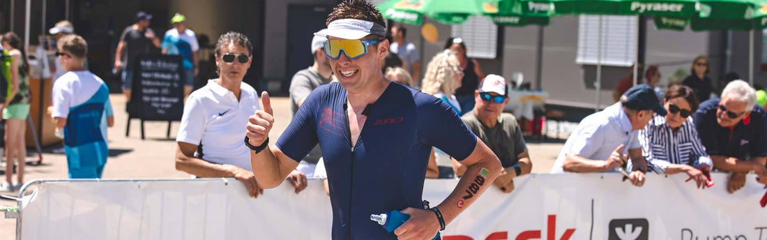 Triathlete in a dark blue suit with a white visor and mirrored sunglasses smiles and gives a thumbs up as spectators watch behind barriers