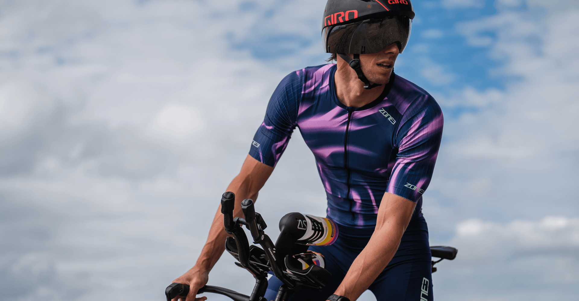 Cyclist in a purple Zone3 kit and Giro helmet rides a bike against a cloudy sky
