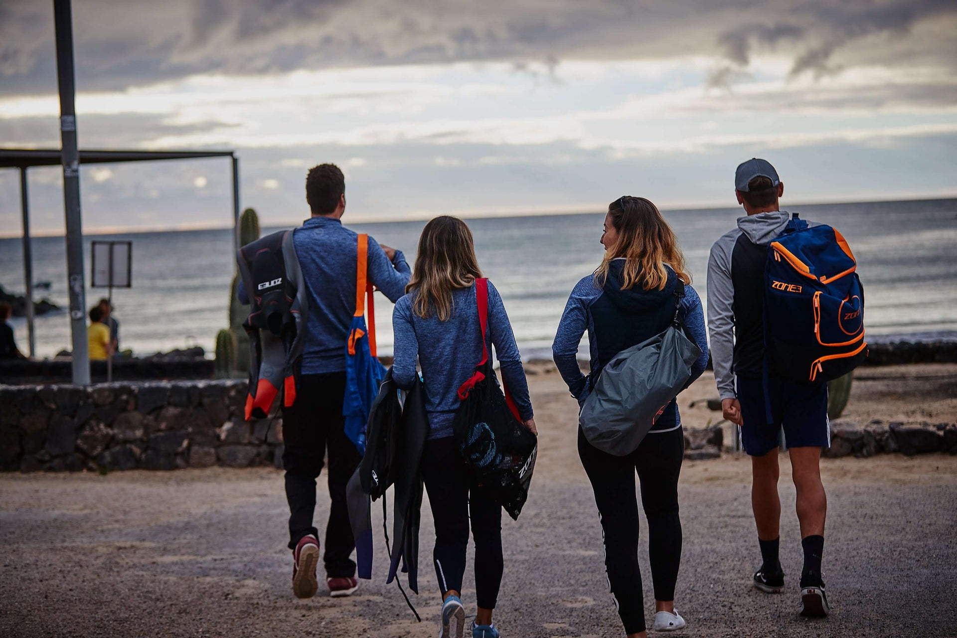 Four travelers with backpacks stand at the waterline, backs to camera, looking out to the sea on a cloudy day.