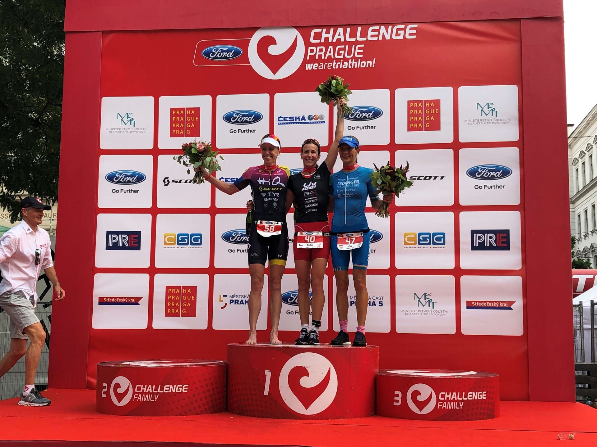 Three athletes stand on a red winners podium at the Challenge Prague awards ceremony, holding bouquets.