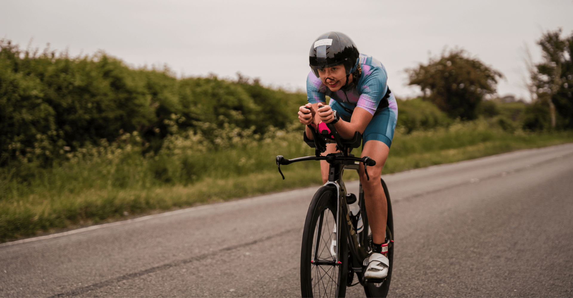 Female cyclist in a teal and pink trisuit leaning forward on a road bike on a rural road.