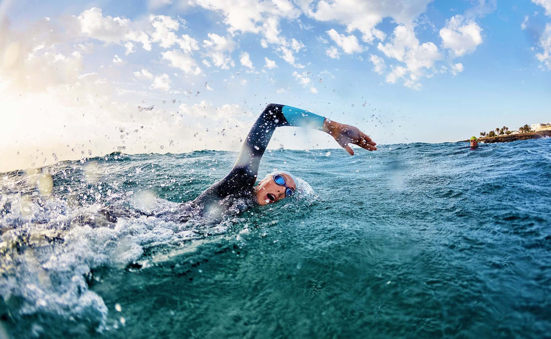 Open water swimmer in a blue wetsuit glides through turquoise water with a sunny coastline and palm trees in the distance.