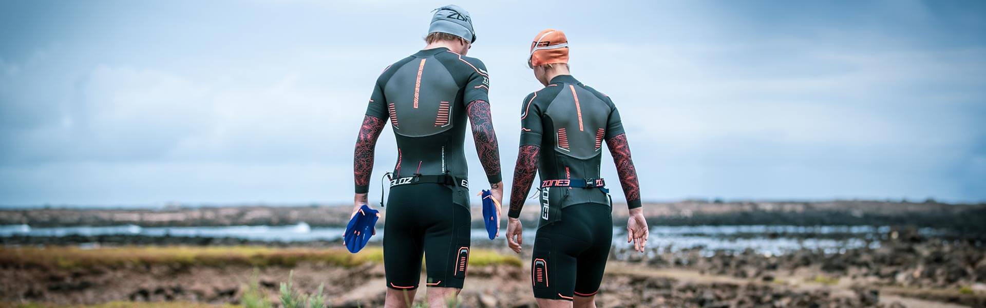 Two athletes in Zone3 wetsuits stand on a rocky coastline with blue swimrun fins in hand.