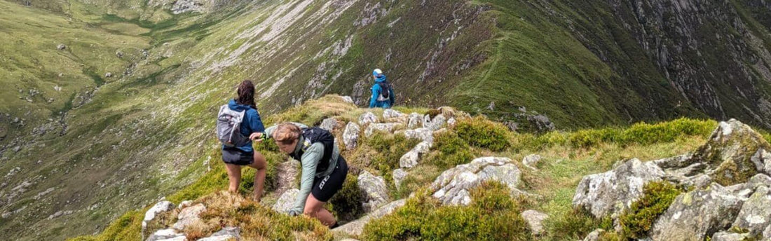 Hikers with backpacks traverse a rocky mountain path surrounded by green slopes.