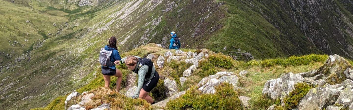 Hikers with backpacks traverse a rocky mountain path surrounded by green slopes.