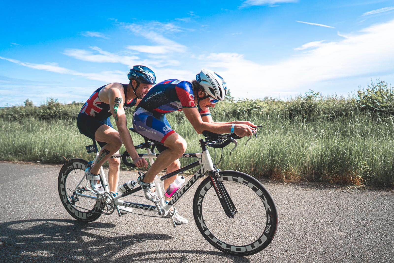 British Paratriathlon Championships competitors ride a tandem bike along a coastal road in aero suits.