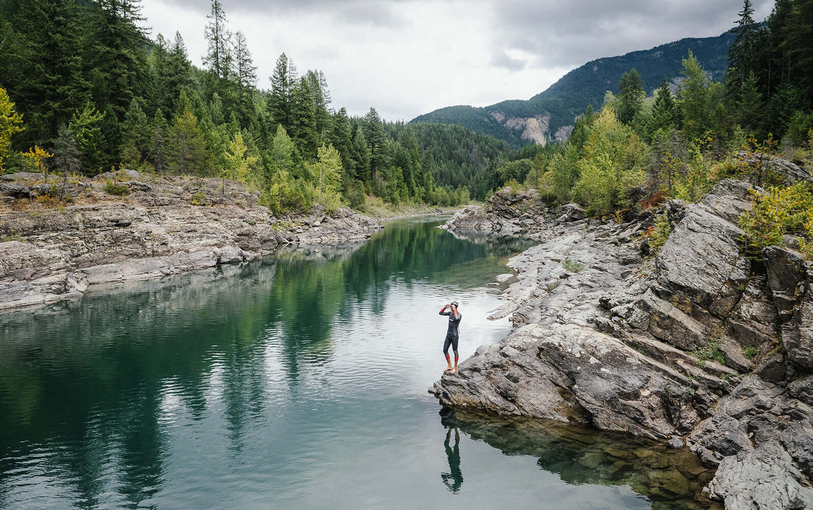 Triathlete in a wetsuit stands on a rocky riverbank beside a calm emerald river flanked by forested cliffs.