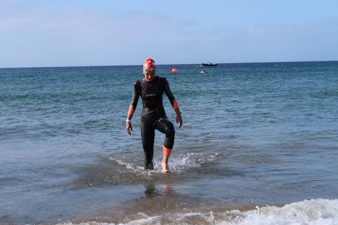 Ironman Lanzarote swimmer in a black wetsuit and red cap emerging from the sea onto the shore.