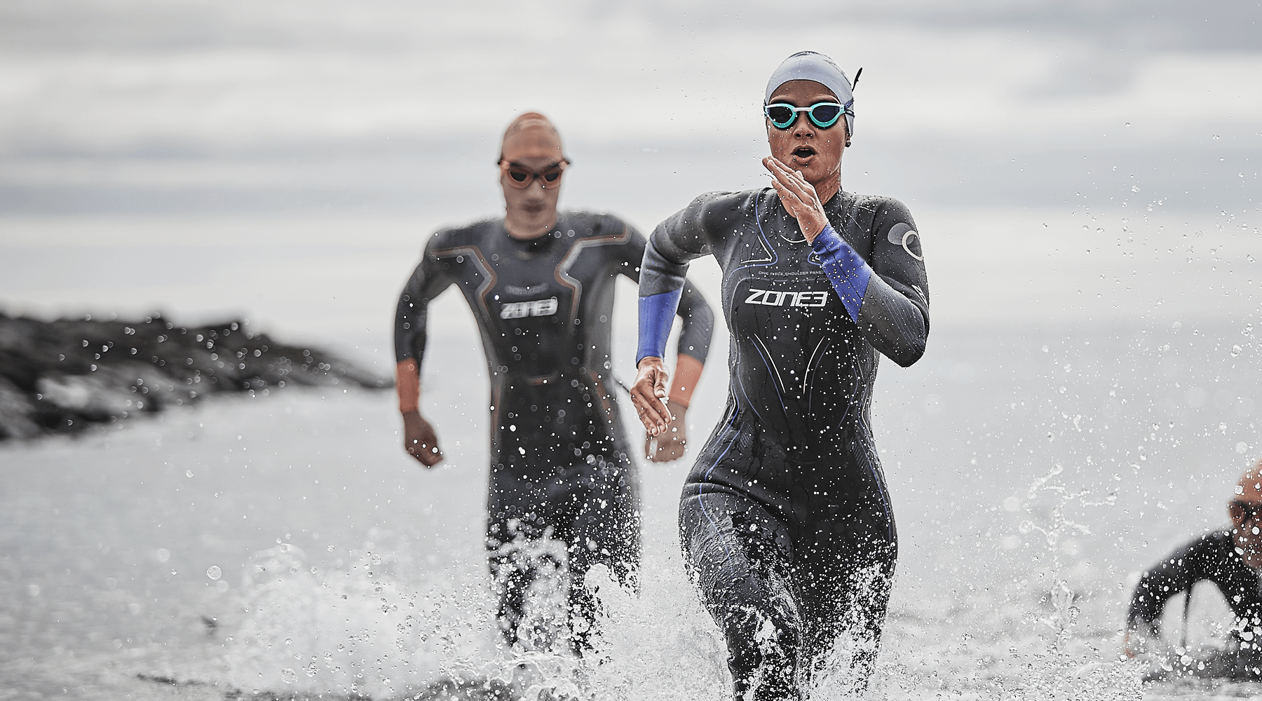 Two triathletes in wetsuits sprint from the water as waves spray around them.