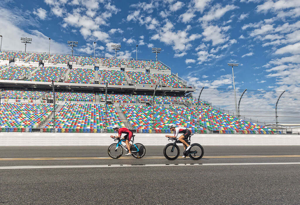 Two cyclists ride on a straight track in front of a colorful stadium with multicolored seating under a bright blue sky.