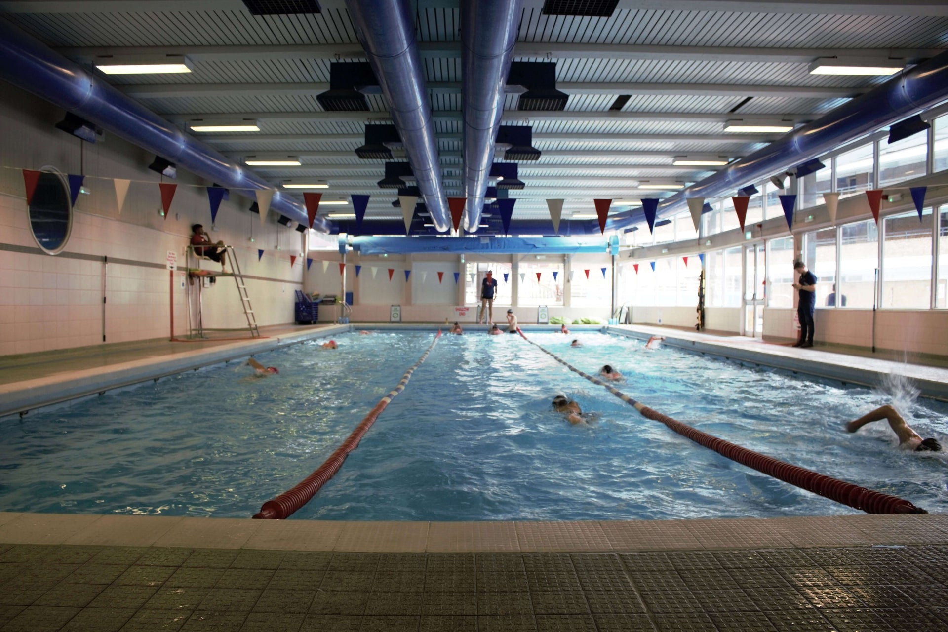 Indoor swimming pool with blue overhead pipes, red white blue pennant banners, and swimmers racing in lanes