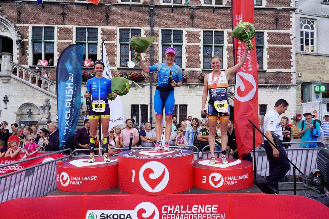 Three female cyclists on the winners podium holding bouquets, in front of a brick building and cheering crowd.