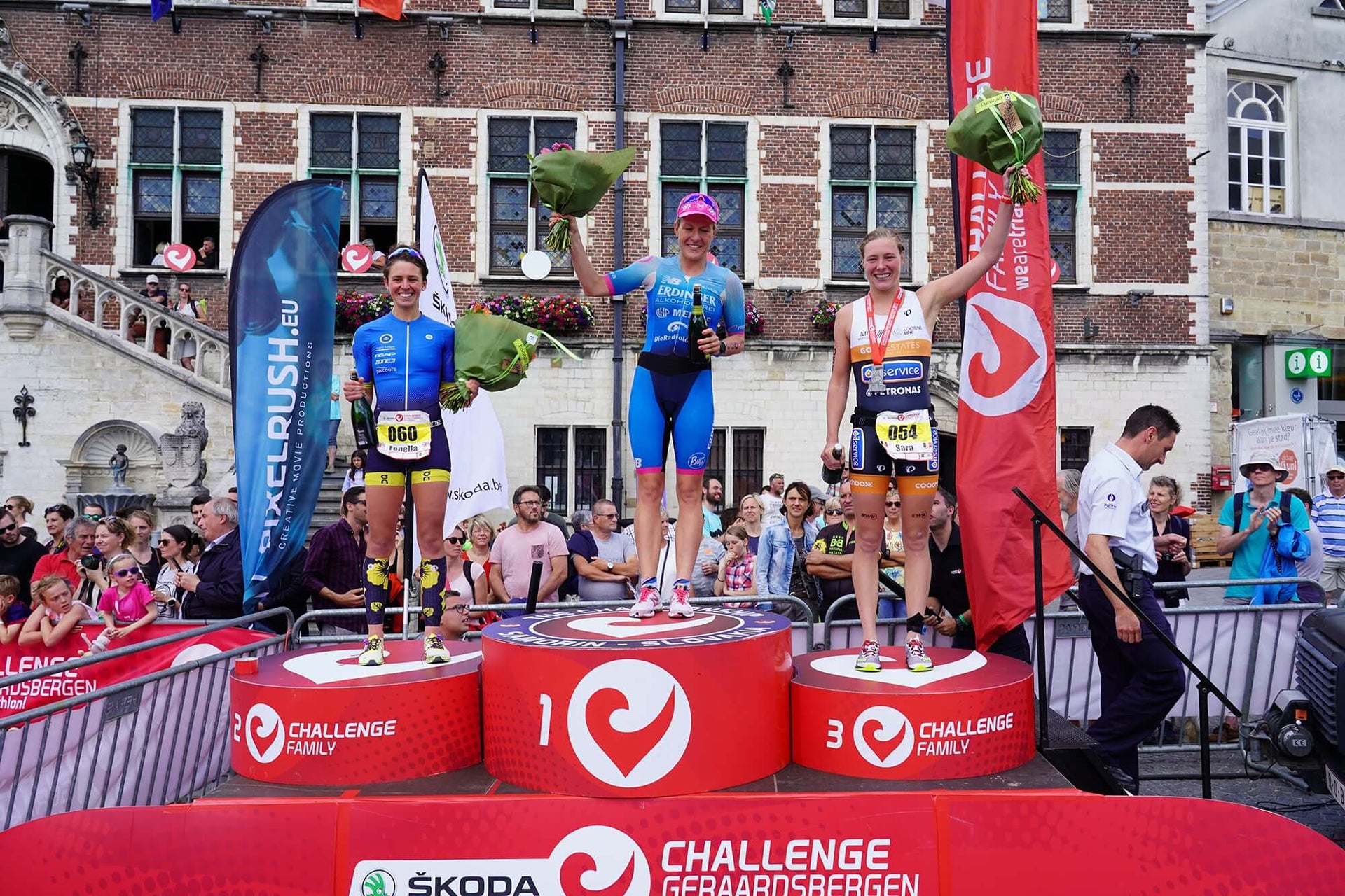 Three female cyclists on the winners podium holding bouquets, in front of a brick building and cheering crowd.