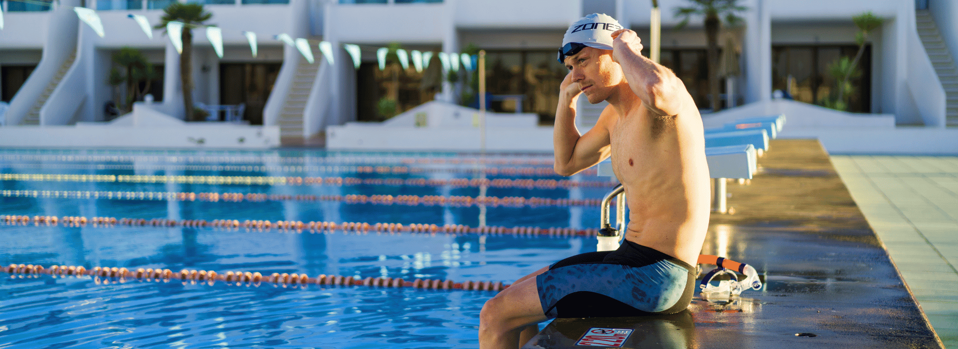 casper stornes sitting on the side of a swimming pool wearing jammers, adjusting swim cap