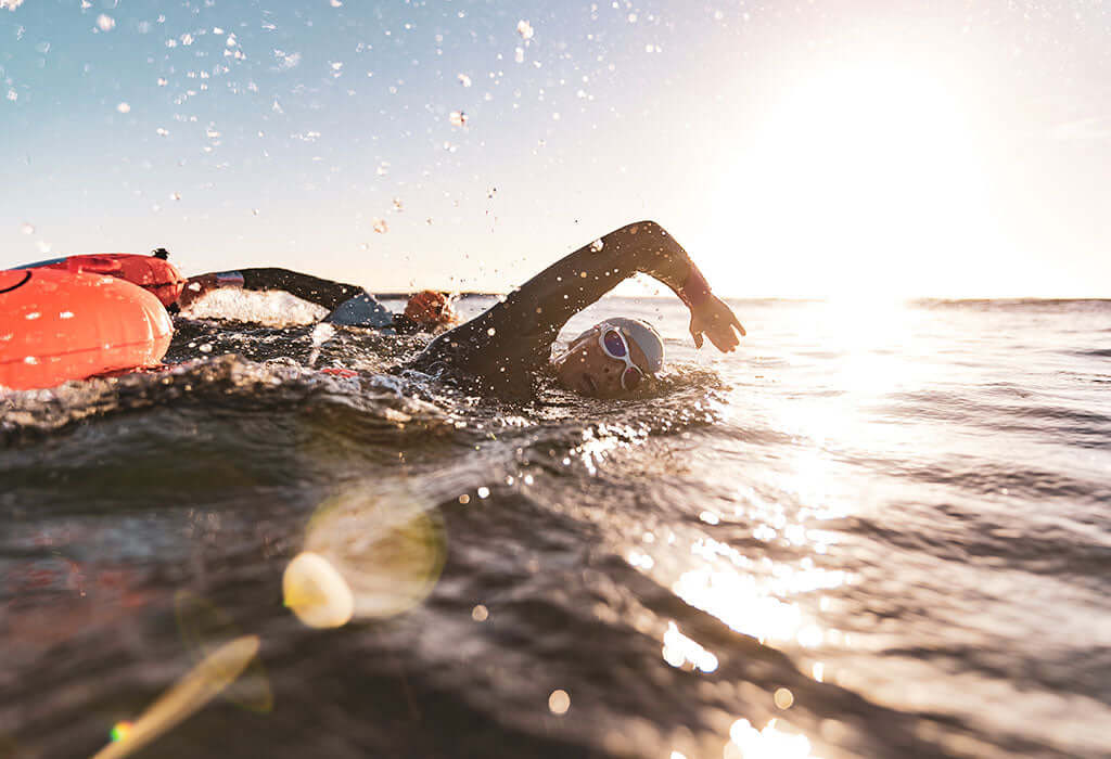 Swimmer in a wetsuit and pink cap swims at sunset with an orange buoy in the background.