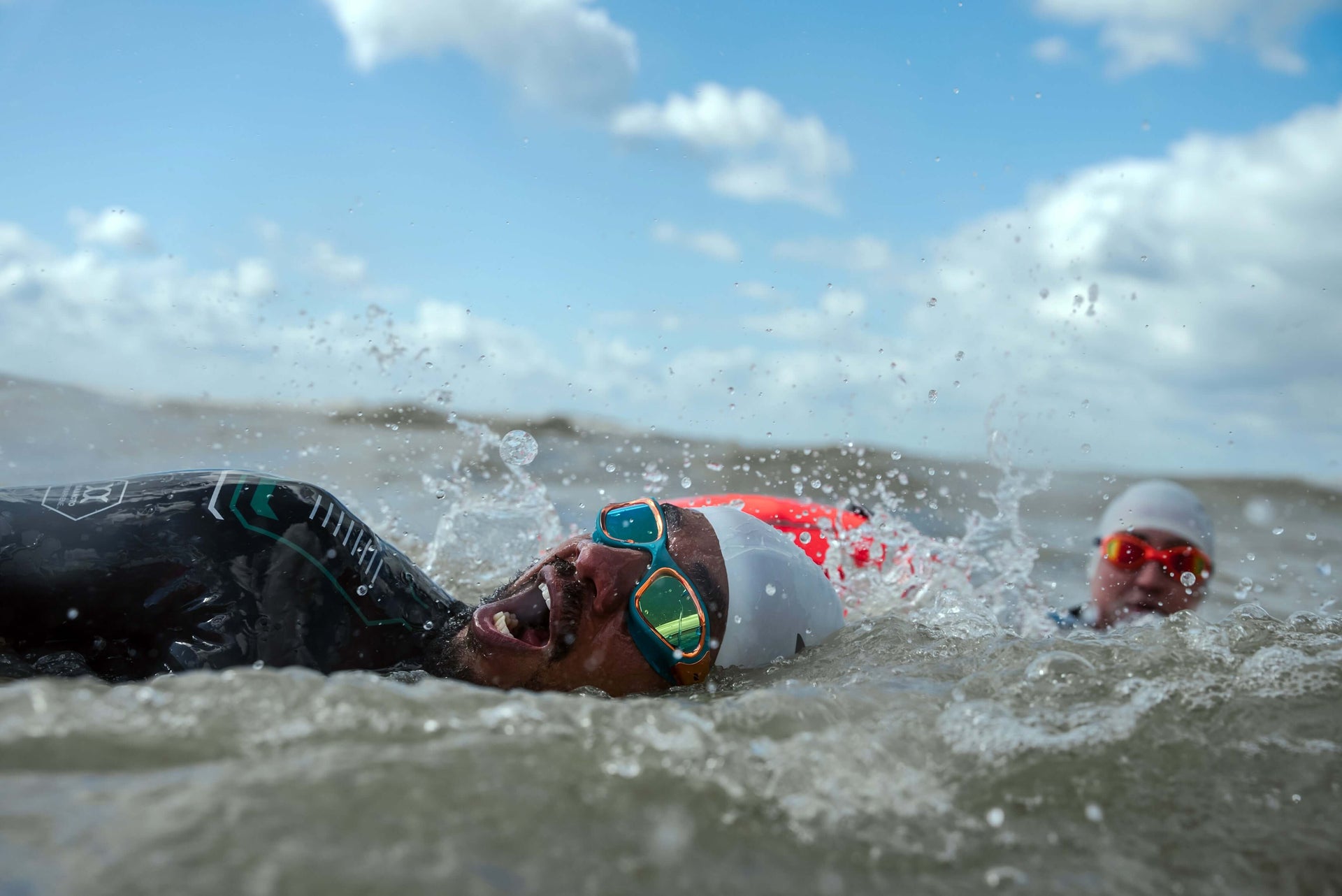 Open water swimmers in wetsuits and goggles swim in choppy water near a sandy shore