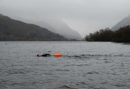 Swimmer in a wetsuit with an orange buoy glides through a misty lake backed by mountains.
