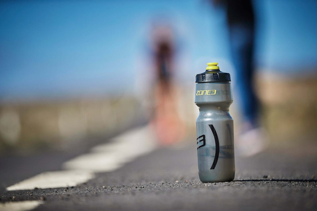 Close-up of a gray Zone3 water bottle with a yellow cap on a road, blurred cyclist in the background