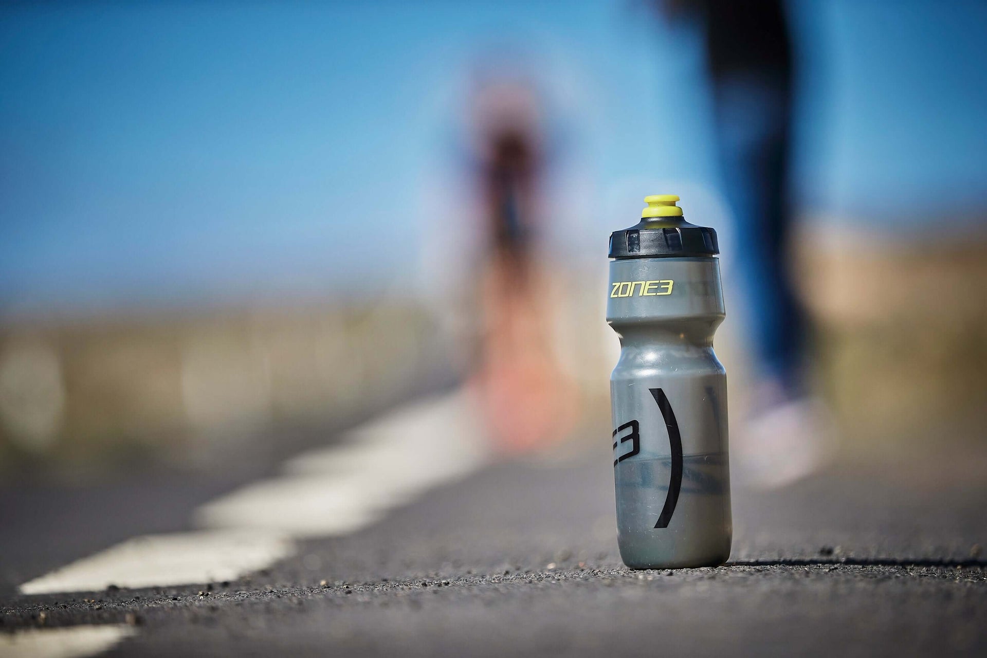 Close-up of a gray Zone3 water bottle with a yellow cap on a road, blurred cyclist in the background