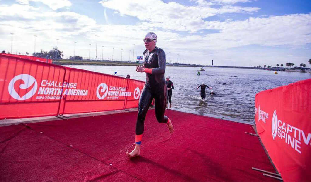 Triathlete sprinting toward the finish line on a red carpet with water in the background and Challenge North America banners.