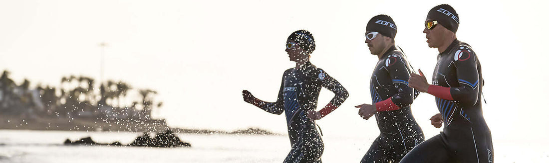 Three Zone3 triathletes in wetsuits run along the sunlit shoreline after a swim