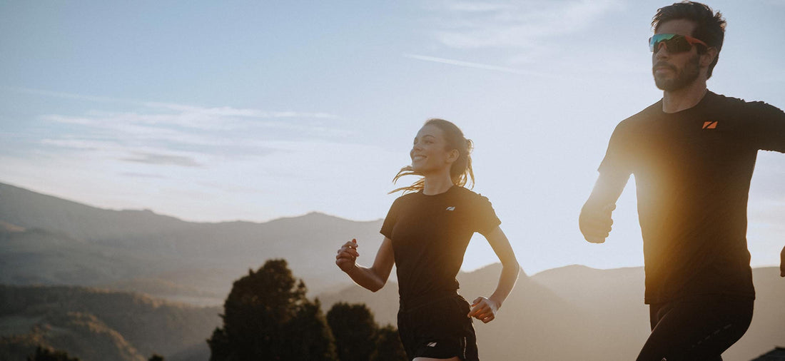 Two runners in Zone3 athletic gear sprint along a sunlit hillside trail with distant mountains.
